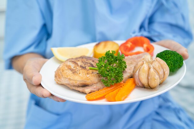 Paciente idosa asiática ou idosa tomando café da manhã e comida saudável vegetal com esperança e feliz enquanto está sentado e com fome na cama no hospital.