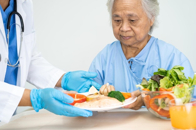 Paciente idosa asiática ou idosa tomando café da manhã e comida saudável vegetal com esperança e feliz enquanto está sentado e com fome na cama no hospital