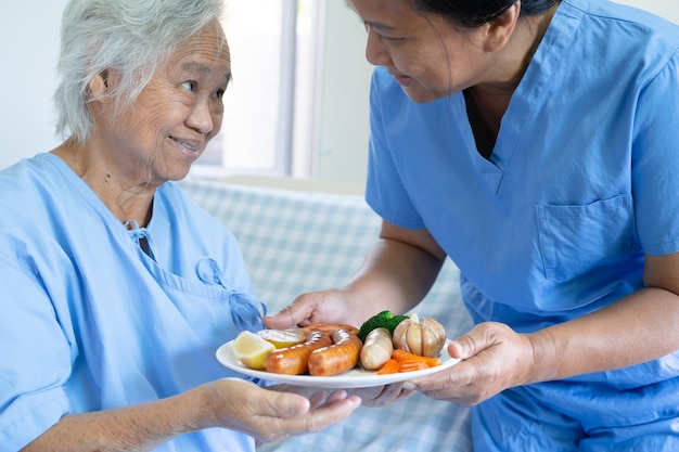 Paciente idosa asiática ou idosa tomando café da manhã e comida saudável vegetal com esperança e feliz enquanto está sentado e com fome na cama no hospital