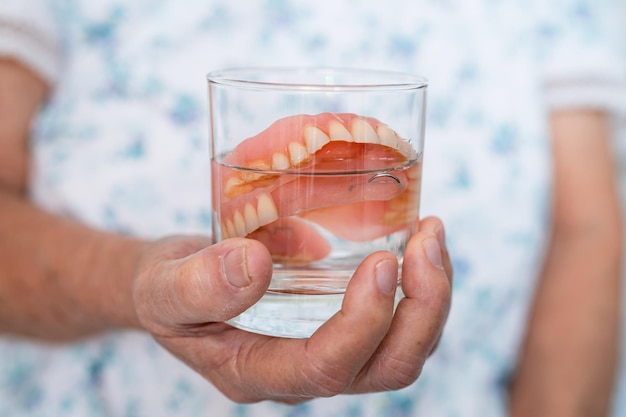 Paciente idosa asiática segurando e lavando dentadura em vidro de limpeza de água para boa mastigação