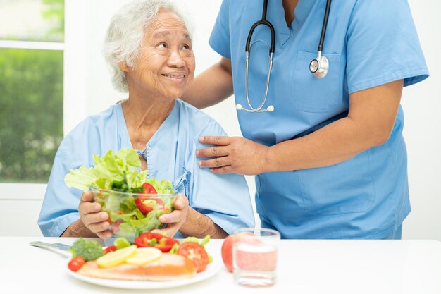 Paciente idosa asiática tomando café da manhã de filé de salmão com alimentos vegetais saudáveis no hospital.