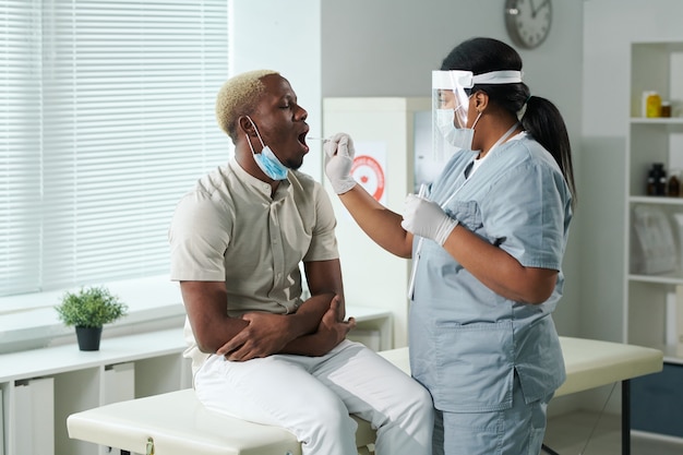Paciente jovem africano do sexo masculino com boca aberta sendo testado para cobiça por enfermeira mestiça, usando roupa de trabalho de proteção segurando cotonete oral
