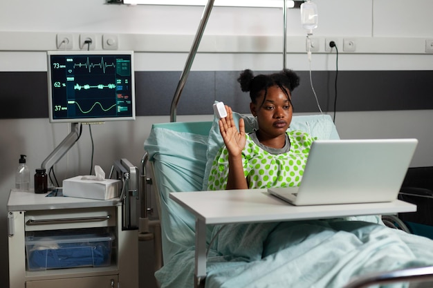 Paciente jovem afro-americano, doente, deitado na cama, cumprimentando um amigo remoto, explicando a experiência de doença durante a conferência de videochamada online na enfermaria do hospital. chamada de telecomunicação
