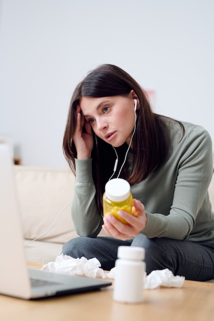 Paciente jovem doente do sexo feminino em casa segurando um frasco de comprimidos enquanto está sentado no sofá à mesa em frente ao laptop e consultando um médico online
