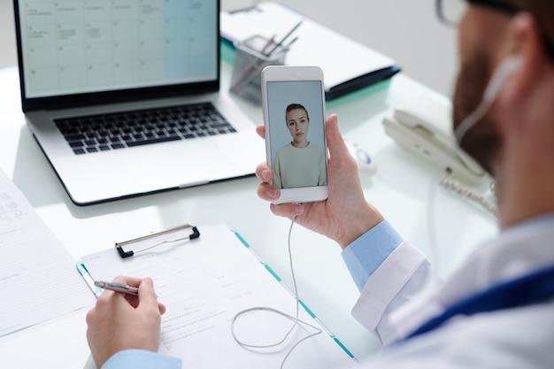 Paciente jovem na tela do smartphone olhando para o médico com documento médico fazendo anotações e dando recomendações médicas