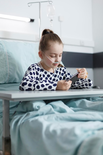 Paciente joven enfermo jugando en el teléfono inteligente mientras descansa en la cama en la habitación del hospital pediátrico. niña enferma y alegre que disfruta del entretenimiento del teléfono móvil mientras se sienta en la sala de recuperación de la clínica.