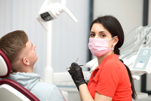 El paciente joven feliz está sentado en la silla del médico dentista en la clínica del consultorio dental y la hermosa doctora con máscara médica en la cara y los guantes Visitando al dentista para revisar los dientes