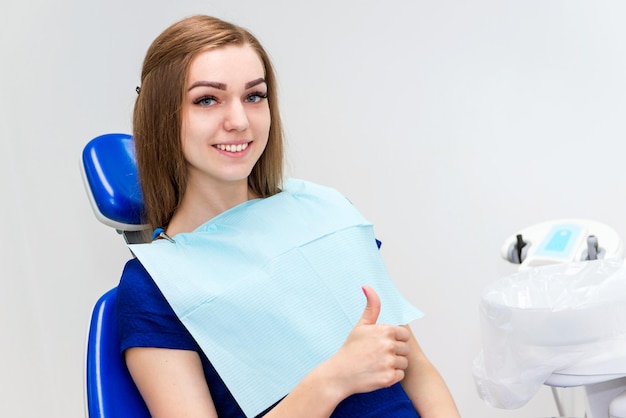 La paciente joven sonríe y muestra el pulgar hacia arriba en la silla del médico en el consultorio dental