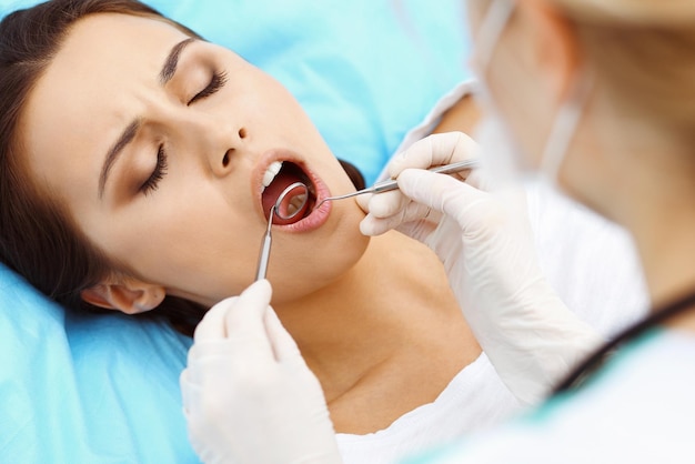 Paciente joven visitando el consultorio del dentista. Hermosa mujer con dientes blancos rectos sanos sentada en el sillón dental con la boca abierta durante el chequeo oral mientras el médico trabaja en los dientes. Clínica dental,