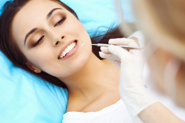 Paciente joven visitando el consultorio del dentista. Hermosa mujer con dientes blancos rectos sanos sentada en el sillón dental con la boca abierta durante el chequeo oral mientras el médico trabaja en los dientes. Clínica dental,