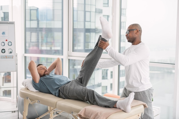 Paciente masculino. Agradable hombre agradable acostado en el sillón médico y levantando la pierna