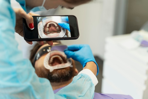 Paciente masculino com a boca aberta sentado na cadeira dentária, dentista segurando um telefone celular tirando uma foto