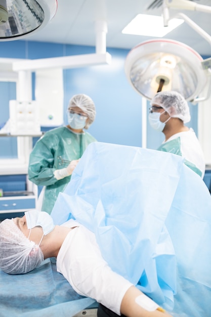 Paciente masculino joven en máscara protectora y sombreros acostado en la mesa de operaciones mientras dos cirujanos en ropa de trabajo se preparan para la operación