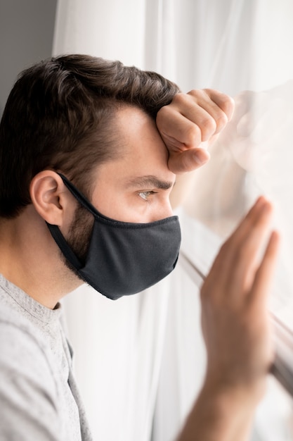 Paciente masculino joven triste en máscara facial mirando por la ventana y apoyándose en él en el hospital