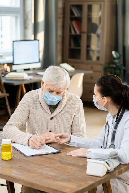 Paciente masculino sênior doente usando máscara e roupa casual, assinando papel médico enquanto está sentado à mesa em frente a uma jovem médica na sala de estar