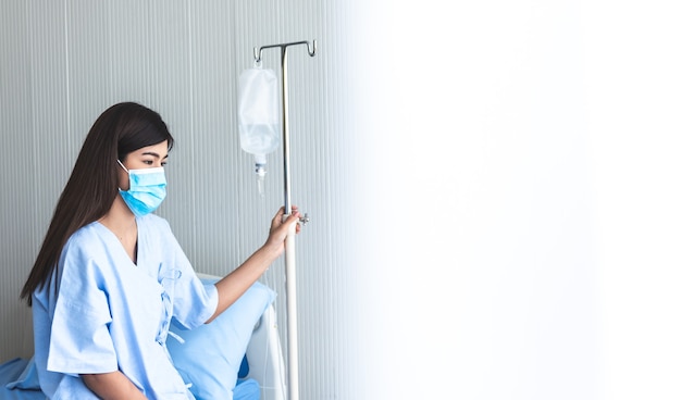 Paciente mulher asiática usando máscaras cirúrgicas, sentada na cama do paciente e segurando infusões de soro fisiológico