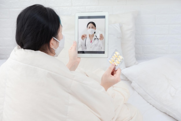 Paciente mulher examina a temperatura corporal durante a consulta com um médico inteligente. Mulher doente na cama videochamada com o médico usando o tablet. Conceito de telemedicina para isolamento domiciliar e aconselhamento on-line de pacientes