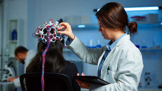 Paciente mulher usando fone de ouvido eeg performant, escaneando a atividade elétrica do cérebro no laboratório de pesquisa neurológica, enquanto o pesquisador médico o ajusta, examinando o sistema nervoso, digitando no tablet.