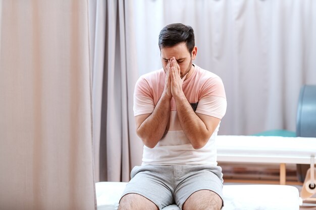 Paciente nervoso, sentado na cama do hospital e orando por bons resultados.