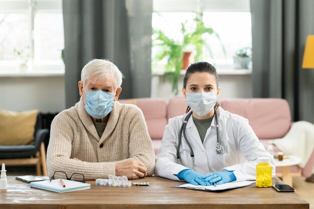 Paciente sênior do sexo masculino doente e jovem morena médica com máscaras protetoras, sentados à mesa um ao lado do outro, olhando para vocês