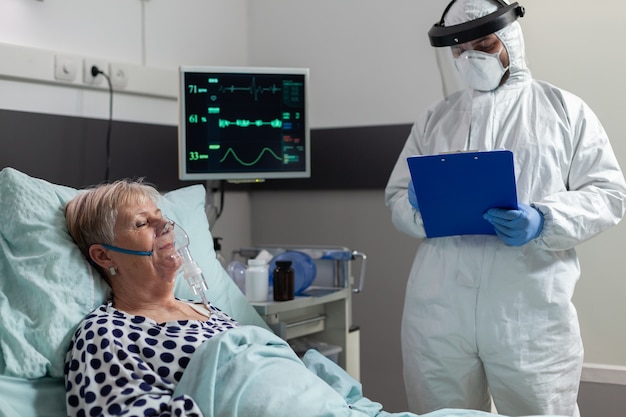 Paciente sênior doente recebendo medicamento intravenoso de uma bolsa de gotejamento iv, deitado na cama, inspira e expira através de máscara de oxigênio, durante a pandemia de coronavírus. médico vestindo terno ppe.