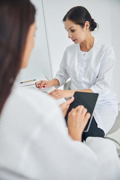 Paciente de sexo femenino de pelo oscuro con una tableta sentado por un médico escribiendo en su computadora portátil