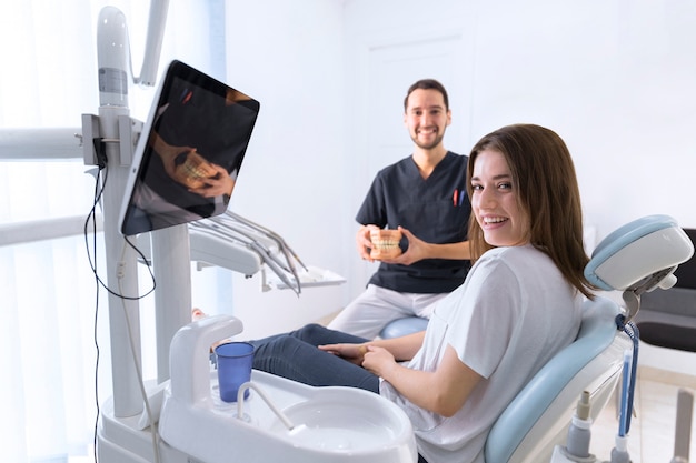 Paciente sonriente sentado en la silla en la clínica delante de dentista sosteniendo el modelo de los dientes