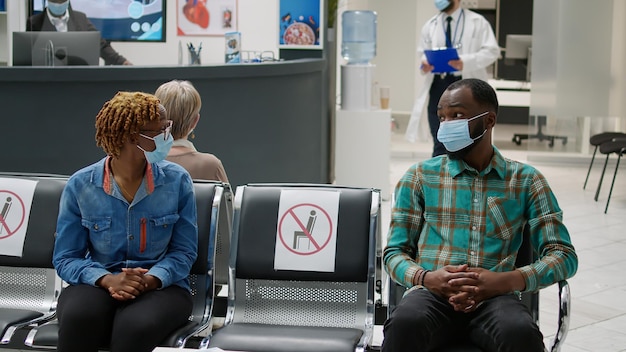 Pacientes afroamericanos con mascarilla hablando en la sala de espera, preparándose para comenzar el examen de control con el médico durante la pandemia de covid 19. Hombre y mujer sentados en la recepción del hospital.