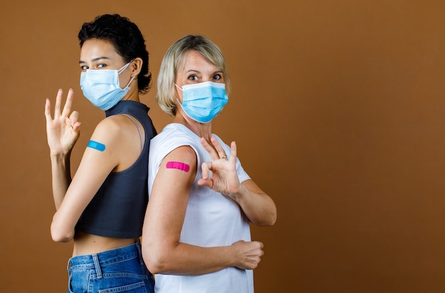 Las pacientes caucásicas usan una mascarilla de pie, miran a la cámara, se apoyan entre sí y muestran el signo de la mano bien o bien en el yeso de colores juntos después de la vacunación frente a un fondo marrón.