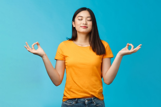 Pacífica encantadora relaxada menina asiática budista meditando, respirando, inalar ar fresco, prática de ioga, fechar os olhos sorrindo aliviado, ficar em pose de nirvana de lótus, alcançar o zen, fundo azul de pé.