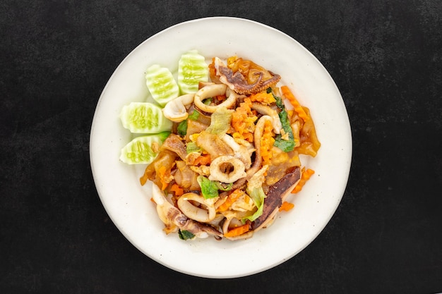 Pad See Ew Pla Muek, comida tailandesa, fideos de salsa de soja salteados con calamares, huevo, lechuga, zanahoria, servido con pepino sobre fondo de textura de tono oscuro, vista superior