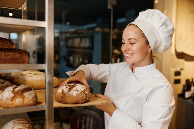 Padeiro de mulher bonita de uniforme verificando pão acabado de cozer na prateleira da bandeja na cozinha moderna da padaria
