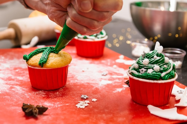 Padeiro fazendo guloseimas de Natal Cupcakes em forma de árvore de Natal As mãos espremem um creme saboroso do saco de confeiteiro em cima dos bolos do festival Cobertura de glacê e decoração com bolos e cupcakes de fondant