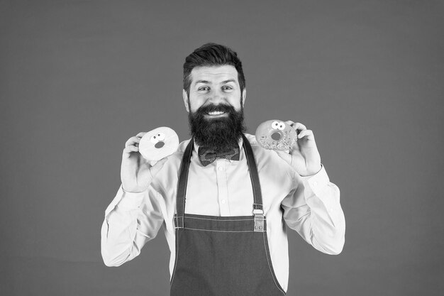 Padeiro habilidoso de entrega de rosquinhas com rosquinha hipster engraçado cozinhar tentação de rosquinha enquanto dieta macho cozinhando padaria homem barbudo no avental do chef garçom brutal na cozinha homem maduro fundo vermelho