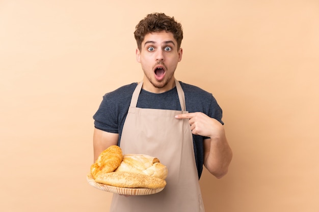 Padeiro masculino segurando uma mesa com vários pães isolados na parede bege com expressão facial de surpresa