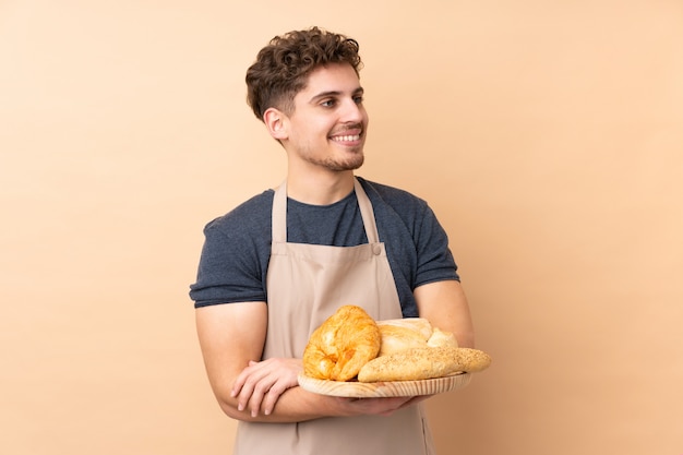 Padeiro masculino segurando uma mesa com vários pães isolados na parede bege, olhando para o lado