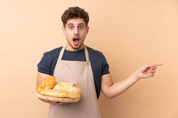 Padeiro masculino segurando uma mesa com vários pães na parede bege surpreendeu e apontando o dedo para o lado