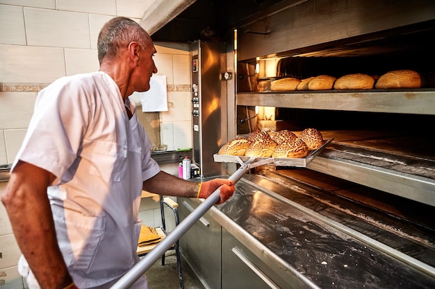 Foto padeiro profissional de uniforme tira um carrinho com pão recém-assado de um forno industrial em uma padaria