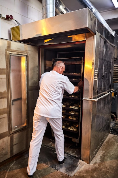 Padeiro profissional em uniforme insere carrinho com decks para assar massa crua para fazer pão em forno industrial em uma padaria