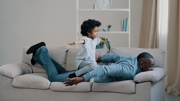 Padre afroamericano adulto acostado boca abajo en el sofá pequeña hija juega se sienta en la espalda de los papás niña