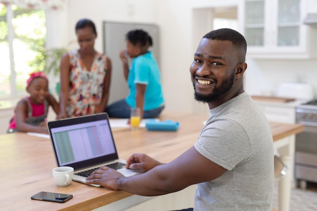 Padre afroamericano que trabaja desde casa usando una computadora portátil con su esposa e hijos haciendo trabajo escolar