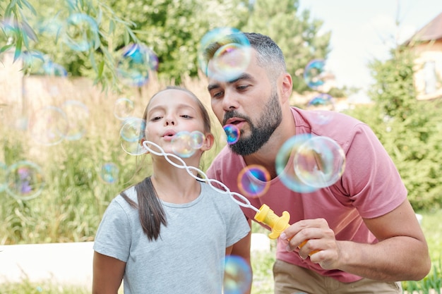 Padre alegre divirtiéndose con su encantadora hija soplando pompas de jabón al aire libre