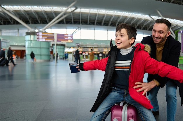 Un padre alegre juega con su adorable hijo haciéndolo rodar en una maleta en la sala de salidas del aeropuerto internacional mientras espera el check-in para el vuelo