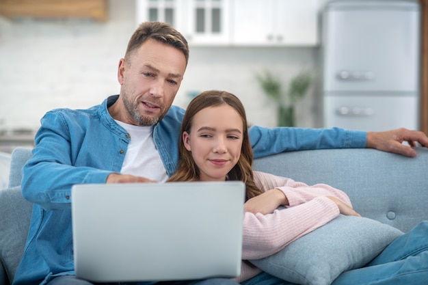 Padre apuntando a la pantalla del portátil e hija sentada en el sofá mirando el portátil de buen humor.