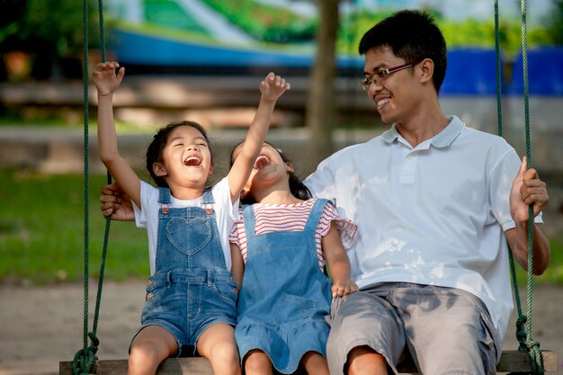 Padre asiático e hija divirtiéndose para montar en columpios juntos en el patio del parque