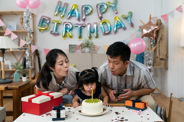 padre asiático, madre e hija se inclinan hacia adelante para soplar velas en el pastel de cumpleaños durante la celebración en casa con globos y guirnaldas de fiesta en el fondo