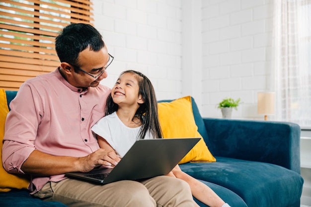Un padre asiático sonriente trabaja desde casa en su portátil mientras su hija aprende en una computadora para el aprendizaje electrónico