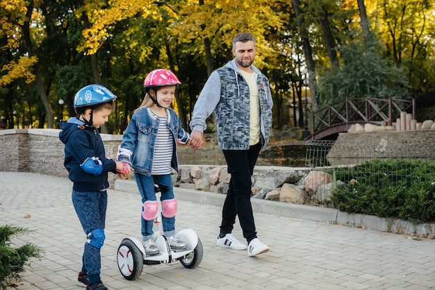 Un padre ayuda y enseña a sus hijos pequeños a montar en Segway en el parque durante la puesta de sol