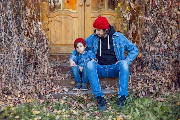 Padre con barba y su hijo con sombreros de punto rojos están sentados en el porche de la casa en Ivy en otoño