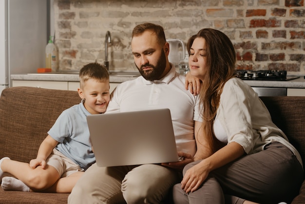 Un padre con barba trabaja de forma remota en una computadora portátil mientras su feliz hijo y su esposa miran la pantalla. Papá está trabajando en línea en una computadora entre parientes en el sofá de casa por la noche